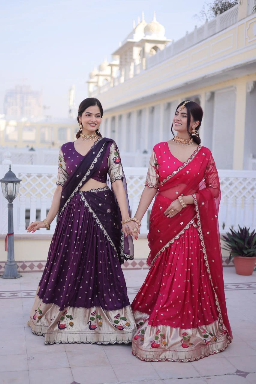 Two women in traditional purple and red sarees standing outdoors with a building in the background.